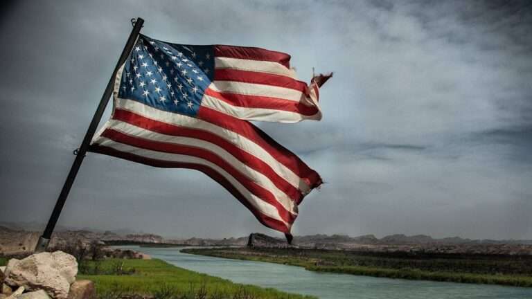 a damaged american flag flying on a pool and a river and clouds can be seen in background.