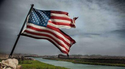 a damaged american flag flying on a pool and a river and clouds can be seen in background.