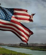 a damaged american flag flying on a pool and a river and clouds can be seen in background.
