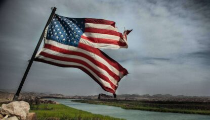 a damaged american flag flying on a pool and a river and clouds can be seen in background.