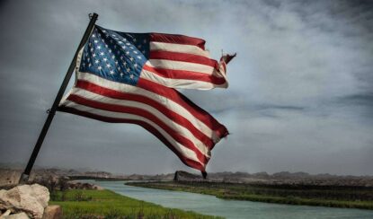 a damaged american flag flying on a pool and a river and clouds can be seen in background.