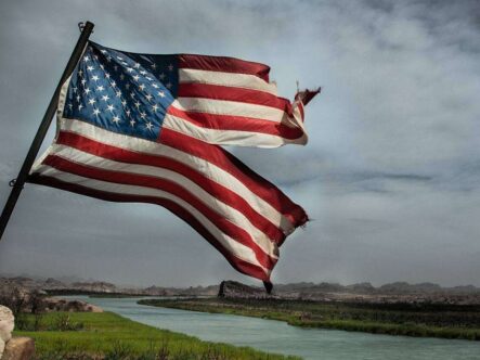 a damaged american flag flying on a pool and a river and clouds can be seen in background.